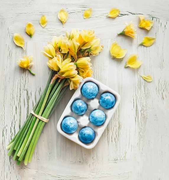 Blue Easter egg in holder with yellow daffodils flowers bunch on white shabby chick background. Top view - Fotoğraf, Görsel
