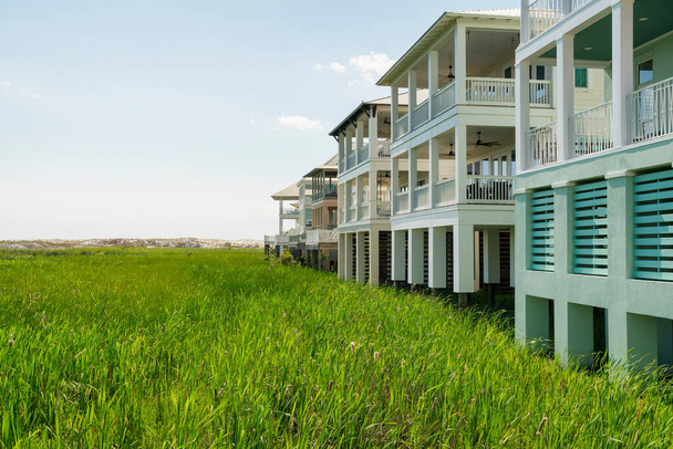 Wysokie trawy na froncie wzniesionych domów z balkonami w Destin na Florydzie. Po lewej stronie jest pole zielonych traw, a po prawej budynki mieszkalne na tle nieba.. - Zdjęcie, obraz