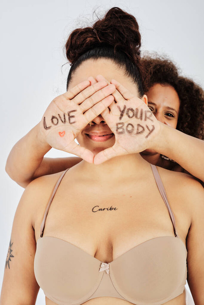 Hispanic female model covering with hands face of ethnic woman in bra while having love your body written on palms - Photo, Image