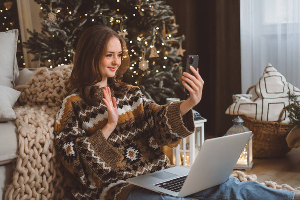 Young beautiful woman with red hair in cozy sweater using mobile smartphone video call to family and friends in room with christmas tree at home. High quality photo - Foto, afbeelding