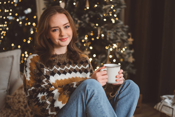 Charming young red-haired woman on the background of a Christmas tree in a cozy living room with a cup of tea. Cheerful red-haired lady puts the ball on the Christmas tree. Womens preparation at home - Foto, afbeelding