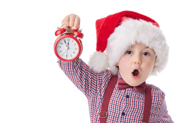 Christmas boy with red alarm clock, smiling little man in red Santa claus hat, tie and suspender posing on white background - Photo, Image