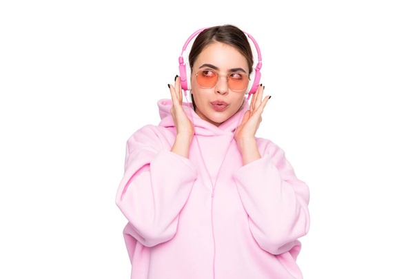 Young woman in casual pink hoodie and glasses listening to music in headphones pouting lips isolated on white. - Foto, imagen