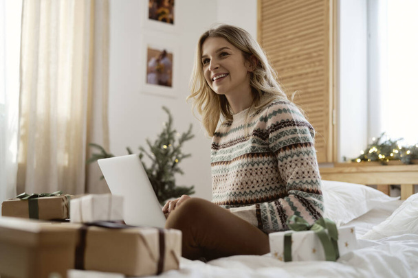 Low angle view of caucasian woman sitting on bed and browsing laptop among Christmas presents - Photo, Image
