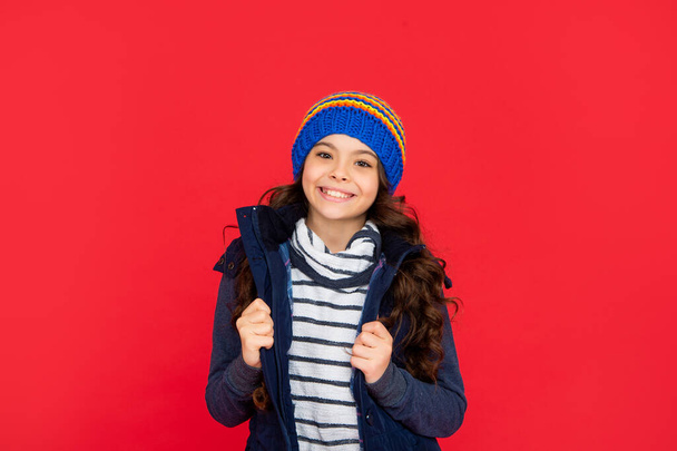 winter fashion. positive kid with curly hair in hat. teen girl on red background. portrait of child wearing warm clothes. express positive emotion. - Photo, image