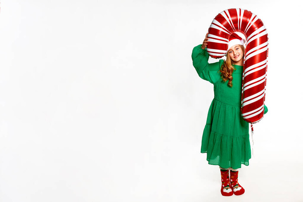 Χριστουγεννιάτικο κορίτσι κρατώντας τεράστιο μπαλόνι από ζαχαροκάλαμο στα χέρια, φορώντας πράσινο φόρεμα, καπέλο santa και κόκκινες ζεστές κάλτσες για τον εορτασμό της Πρωτοχρονιάς κόμμα - Φωτογραφία, εικόνα