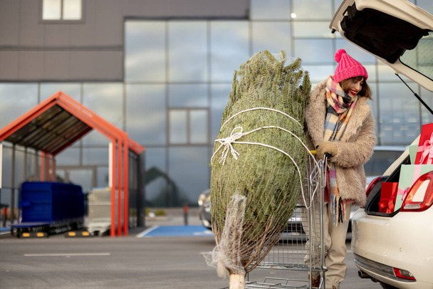 Giovane donna imballaggio albero di Natale e presenta nel bagagliaio auto al parcheggio di un centro commerciale. Ragazza allegra che fa shopping festivo in previsione delle vacanze invernali - Foto, immagini