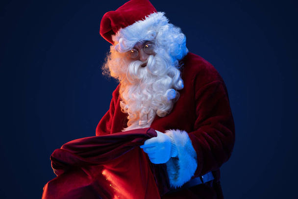 Portrait of old santa claus with glasses and bag with gifts against dark background. - Photo, Image