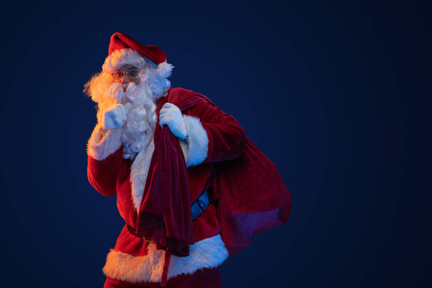 Studio schot van de kerstman gekleed in rode kleren draagtas tegen donkere achtergrond. - Foto, afbeelding
