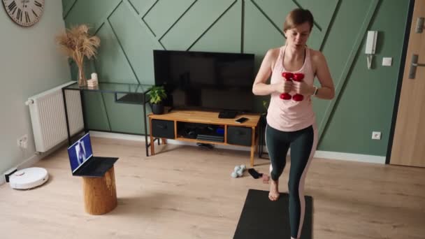 Woman is doing sports exercises in living room. Remote classes with fitness trainer at home, using laptop for video call. Woman in sports clothes does cardio on the floor - Footage, Video