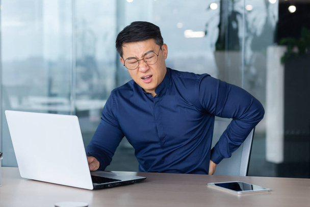 Sick overworked man at work, businessman has severe back pain, asian man holding hand on side massaging muscle, boss in shirt and glasses at work inside office using laptop. - Фото, зображення