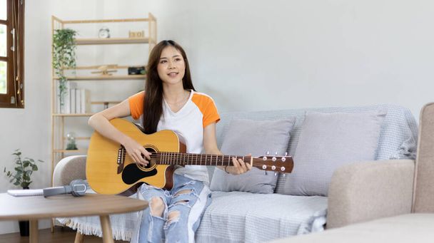 Azjatycka artystka grająca na gitarze i śpiewająca szczęśliwie w salonie, Relaks z muzykoterapią, Spędzanie wolnego czasu z muzyką, Radość z grania muzyki, Gitara akustyczna. - Zdjęcie, obraz