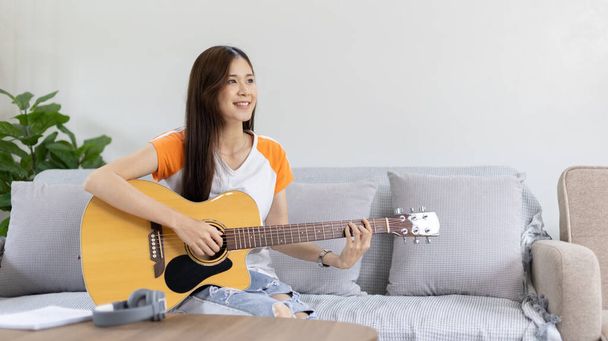 Ασιάτισσα καλλιτέχνιδα που παίζει κιθάρα και τραγουδά χαρούμενα στο σαλόνι, Χαλάρωση με μουσικοθεραπεία, Ξοδεύοντας ελεύθερο χρόνο με μουσική, Χαρά παίζοντας μουσική, Ακουστική κιθάρα. - Φωτογραφία, εικόνα