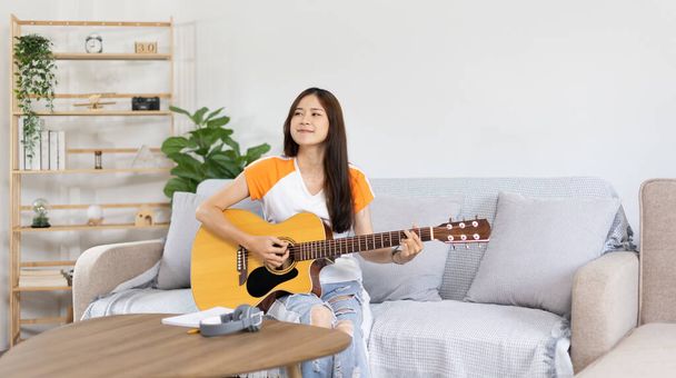 Artista asiatica femminile che suona la chitarra e canta felicemente in salotto, Relax con la musicoterapia, Trascorrere il tempo libero con la musica, Gioia di suonare musica, Chitarra acustica. - Foto, immagini