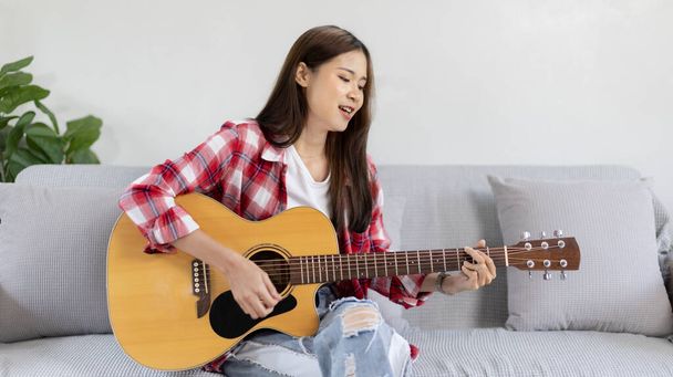 アジアの女性アーティストがギターを弾き、リビングルームで楽しく歌い、音楽療法でリラックスし、音楽と自由な時間を過ごす、音楽を演奏する喜び、アコースティックギター. - 写真・画像