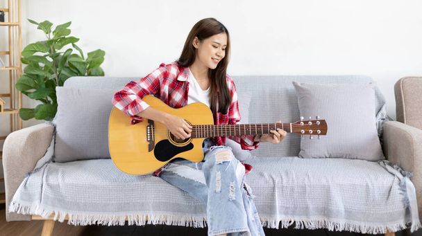 Азиатская исполнительница, играющая на гитаре и счастливо поющая в гостиной, релаксация с музыкальной терапией, свободное время с музыкой, радость от игры, акустическая гитара. - Фото, изображение
