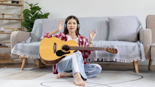 Asiatische Künstlerin spielt Gitarre und singt fröhlich im Wohnzimmer, Entspannung mit Musiktherapie, Freizeit mit Musik verbringen, Freude am Musizieren, Akustikgitarre. - Foto, Bild