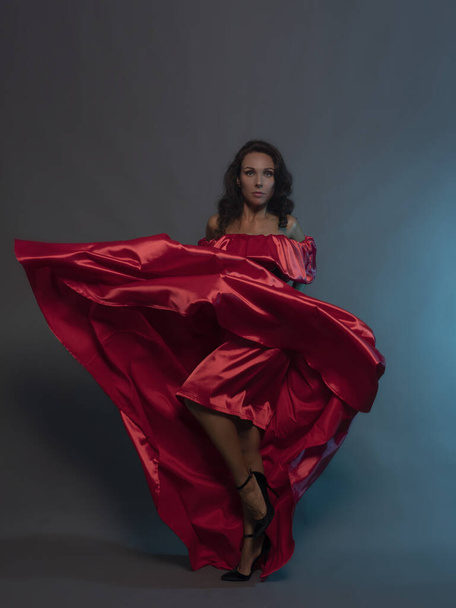 A beautiful brunette in a red flying dress poses in the studio on a gray background.  - Фото, изображение