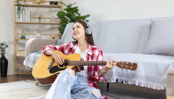 Asiatische Künstlerin spielt Gitarre und singt fröhlich im Wohnzimmer, Entspannung mit Musiktherapie, Freizeit mit Musik verbringen, Freude am Musizieren, Akustikgitarre. - Foto, Bild