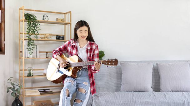 Aasialainen naisartisti soittaa kitaraa ja laulaa iloisesti olohuoneessa, Rentoutuminen musiikkiterapian kanssa, Vapaa-ajan vietto musiikin kanssa, Musiikin soittamisen ilo, Akustinen kitara. - Valokuva, kuva