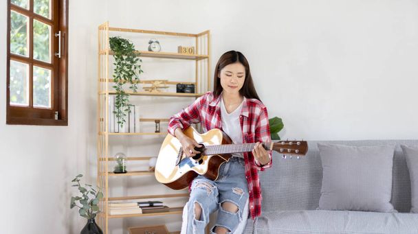 Azjatycka artystka grająca na gitarze i śpiewająca szczęśliwie w salonie, Relaks z muzykoterapią, Spędzanie wolnego czasu z muzyką, Radość z grania muzyki, Gitara akustyczna. - Zdjęcie, obraz