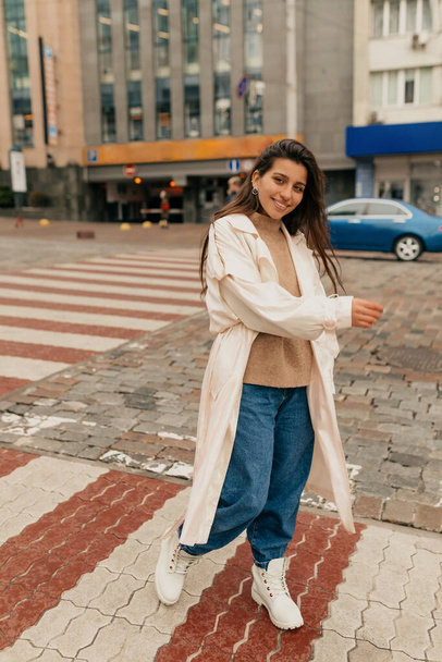 Повнометражне фото привабливої усміхненої жінки з довгим волоссям у білій траншеї, джинсах та светрі, що йдуть по вулиці та посміхаються
  - Фото, зображення