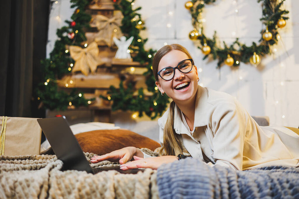Young beautiful woman in cozy home clothes using laptop on bed in room with Christmas tree. Portrait of carefree happy girl student at home in Christmas - Photo, Image