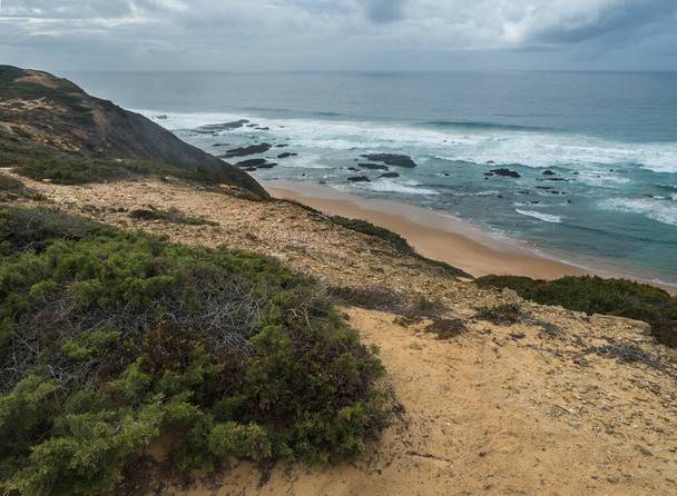 Widok na pustą plażę Praia da Carreagem z falami oceanicznymi, klifami i kamieniami, mokrym złotym piaskiem i zieloną roślinnością na wybrzeżu Rota Vicentina, Odemira, Portugalia. Chmurny jesienny dzień - Zdjęcie, obraz