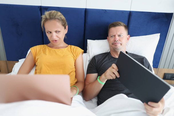 Der eifersüchtige Ehemann verdächtigt seine Frau des Betrugs und blickt auf ihren Computerbildschirm. Familienpaar liegt mit Laptop im Bett. - Foto, Bild