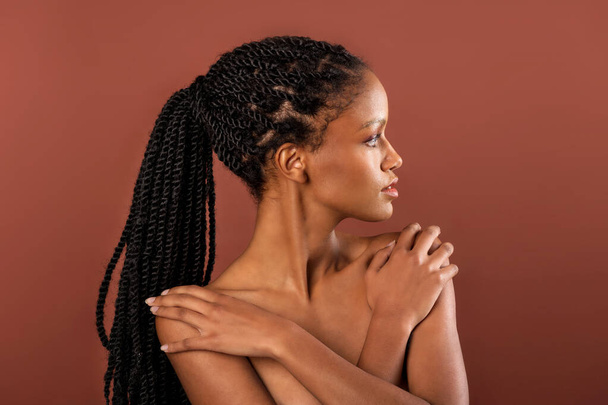 Wunderschöne junge ethnische Frau mit Afro-Zöpfen, die Hände auf nackten Schultern hält und vor braunem Hintergrund wegschaut - Foto, Bild