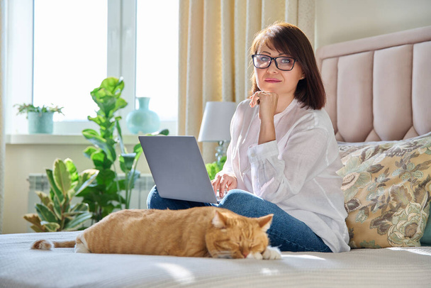 Middle aged woman at home on couch with laptop and cat. Mature 40s female looking at camera, on bed with pet. Work at home, technology, leisure, freelancing, lifestyle, people and animals concept - Zdjęcie, obraz