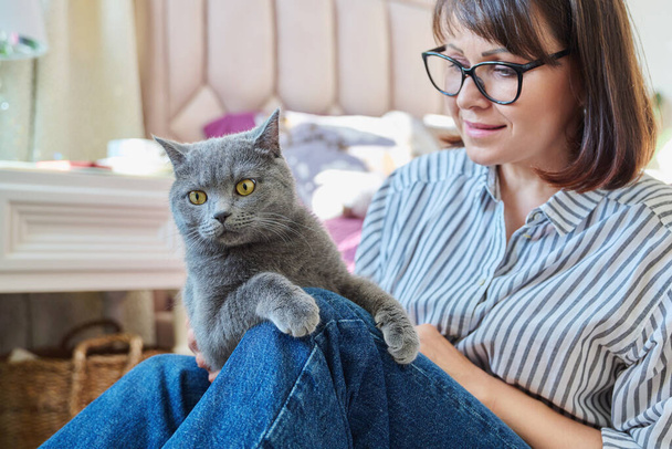 Gray cat in hands of woman at home, cat sitting on knees, woman owner talking touching pet, love friendship, animals people concept - Фото, изображение