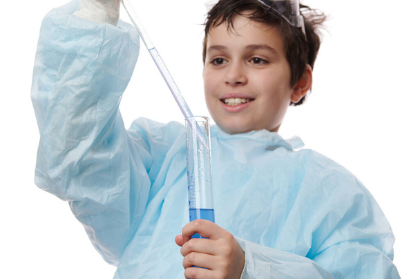 Zeki bir okul çocuğunun yakın plan portresi, genç bir çocuk, genç bir kimyager, pipetini, beyaz arka planda izole edilmiş bir laboratuvar silindirinden mavi kimyasal maddeyle dolduruyor. - Fotoğraf, Görsel