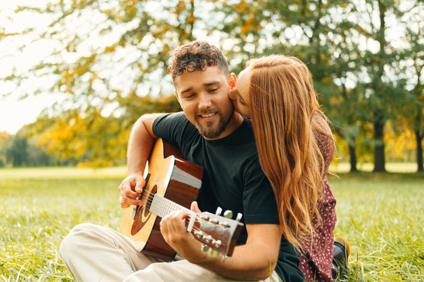 Young woman kisses her man while he plays the guitar sitting on grass in the park. - Photo, Image