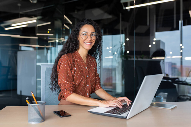 Jovem bela mulher hispânica trabalhando dentro do escritório moderno, mulher de negócios sorrindo e olhando para a câmera no trabalho usando laptop. - Foto, Imagem