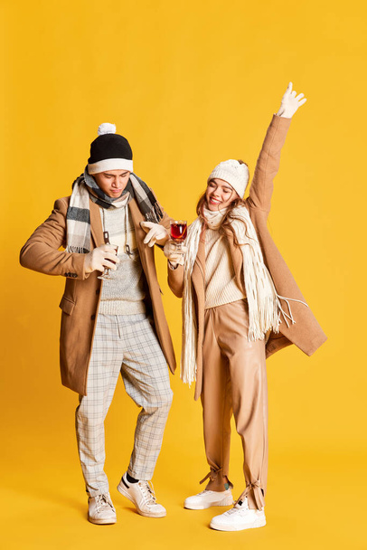 Portrait of stylish, cheerful young man and woman in cozy winter clothes posing, drinking mulled wine isolated over yellow background. Concept of emotions, winter holidays, fashion, lifestyle - Photo, Image