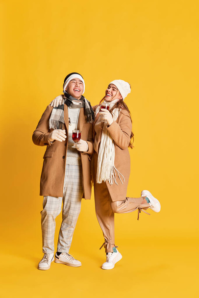 Retrato de jovem e mulher elegante, coupé sorridente em roupas de inverno andando, bebendo vinho quente isolado sobre fundo amarelo. Conceito de emoções, férias de inverno, moda, estilo de vida, diversão - Foto, Imagem