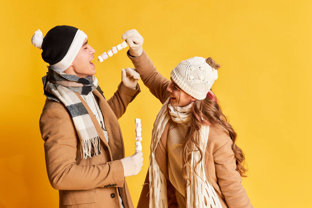 Muotokuva tyylikäs nuori mies ja nainen hymyilee, poseeraa talvivaatteissa, syö vaahtokarkkia eristetty keltaisella taustalla. Käsite tunteita, talvilomat, muoti, elämäntapa, ystävyys - Valokuva, kuva