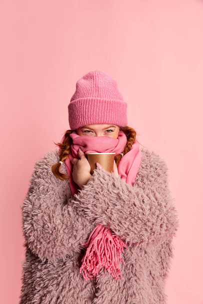 Portrait of young girl in knitted hat, scarf and furry coat warming up with hot drink isolated over pink background. Cozy look. Concept of emotions, winter holidays, fashion, lifestyle, celebration - Fotó, kép