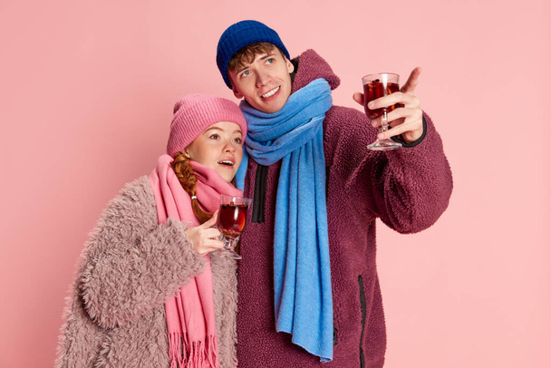 Young man and woman, friends in knitted hat and scarf posing with mulled wine isolated over pink background. Concept of emotions, winter holidays, fashion, lifestyle, celebration, relationship - Photo, Image