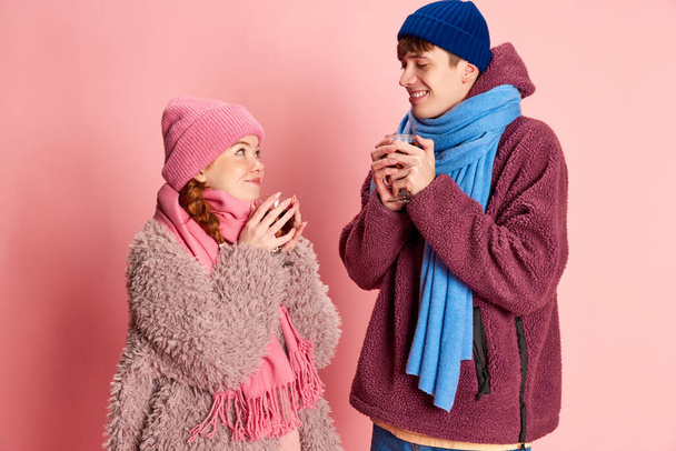 Portrait of young man and woman in knitted hat and scarf posing, warming up with mulled wine isolated over pink background. Concept of emotions, winter holidays, fashion, lifestyle, relationship - Photo, Image
