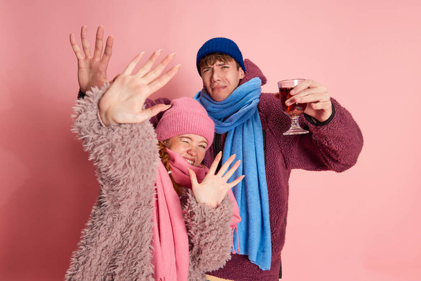 Jeune homme et femme émotif en bonnet tricoté et écharpe posant avec du vin chaud isolé sur fond rose. Concept d'émotions, vacances d'hiver, mode, style de vie, célébration, relation - Photo, image