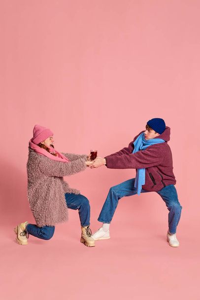 Portrait of young man and woman in knitted hat and scarf posing, fighting for mulled wine glass isolated over pink background. Concept of emotions, winter holidays, fashion, lifestyle, relationship - Photo, Image