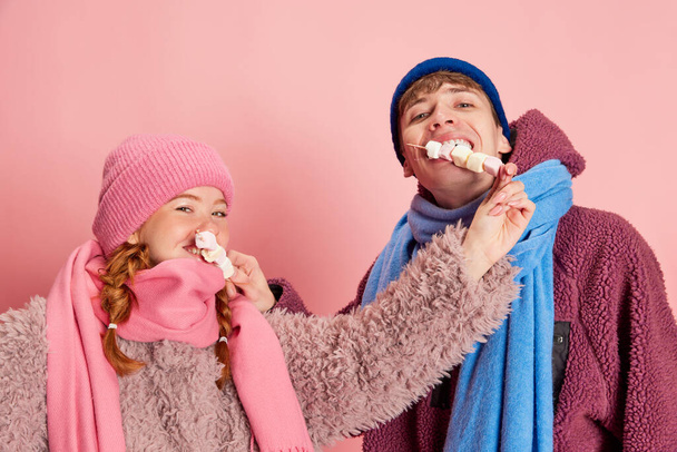Portrait of young man and woman in knitted hat and scarf posing with marshmallow isolated over pink background. Concept of emotions, winter holidays, fashion, lifestyle, celebration, relationship - Foto, Imagen