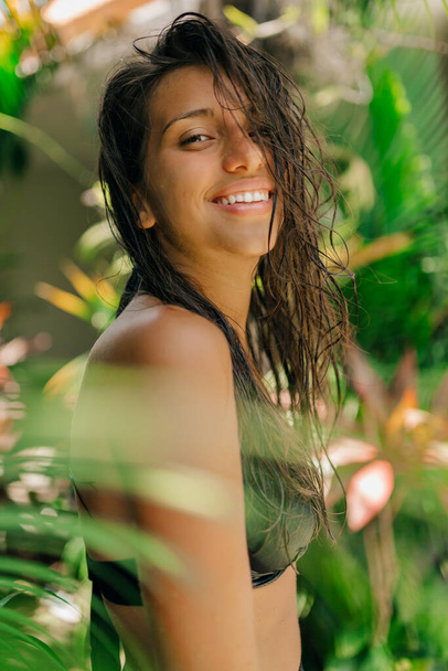 Outdoor portrait of charming brunette woman with adorable smile wearing swim suit posing at camera under palm leaves in sunlight. - 写真・画像