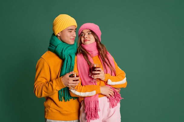 Young man and woman, lovely couple in bright knitted hat and scarf posing with mulled wine isolated over green background. Concept of emotions, winter holidays, fashion, lifestyle, relationship - Valokuva, kuva