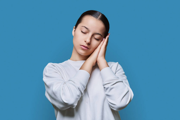 Young woman sleeping with closed eyes, holding hands near ear, sleeping gesture, on blue studio background. Body language, healthy sleep, beauty, lifestyle, youth students concept - Foto, Imagem