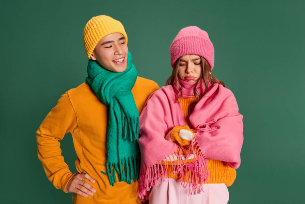 Portret van een jonge man en vrouw met een heldere gebreide hoed en sjaal die geïsoleerd over een groene achtergrond poseren. Een gezicht trekken. Concept van emoties, wintervakantie, mode, levensstijl, feest, relatie - Foto, afbeelding