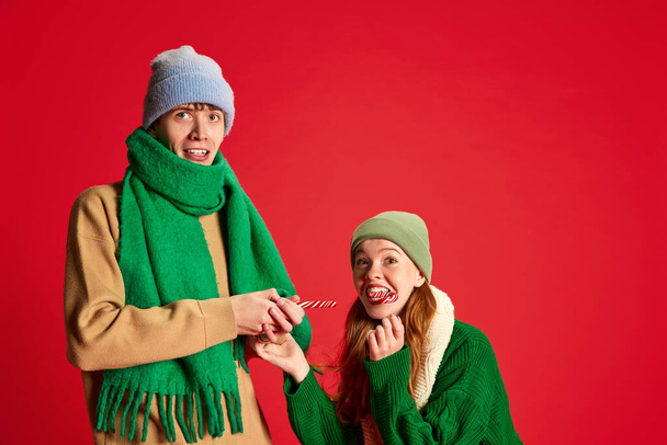 Portrait of young man and woman in green knitted sweater and hat posing isolated on red background. Eating candies. Concept of emotions, winter holidays, fashion, lifestyle, celebration, relationship - Photo, Image