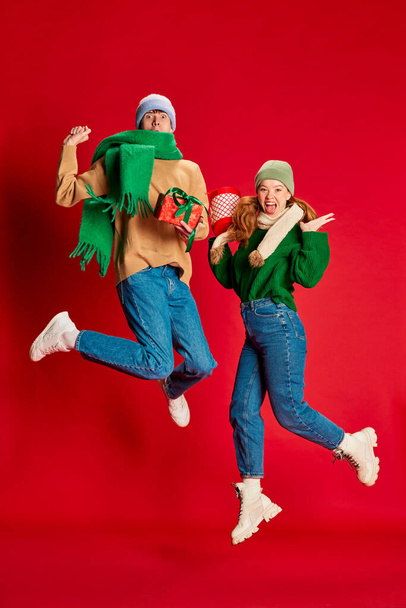 Portrait of young man and woman in green knitted sweater and hat jumping with present boxes isolated over red background. Concept of emotions, winter holidays, fashion, lifestyle, celebration - Fotoğraf, Görsel